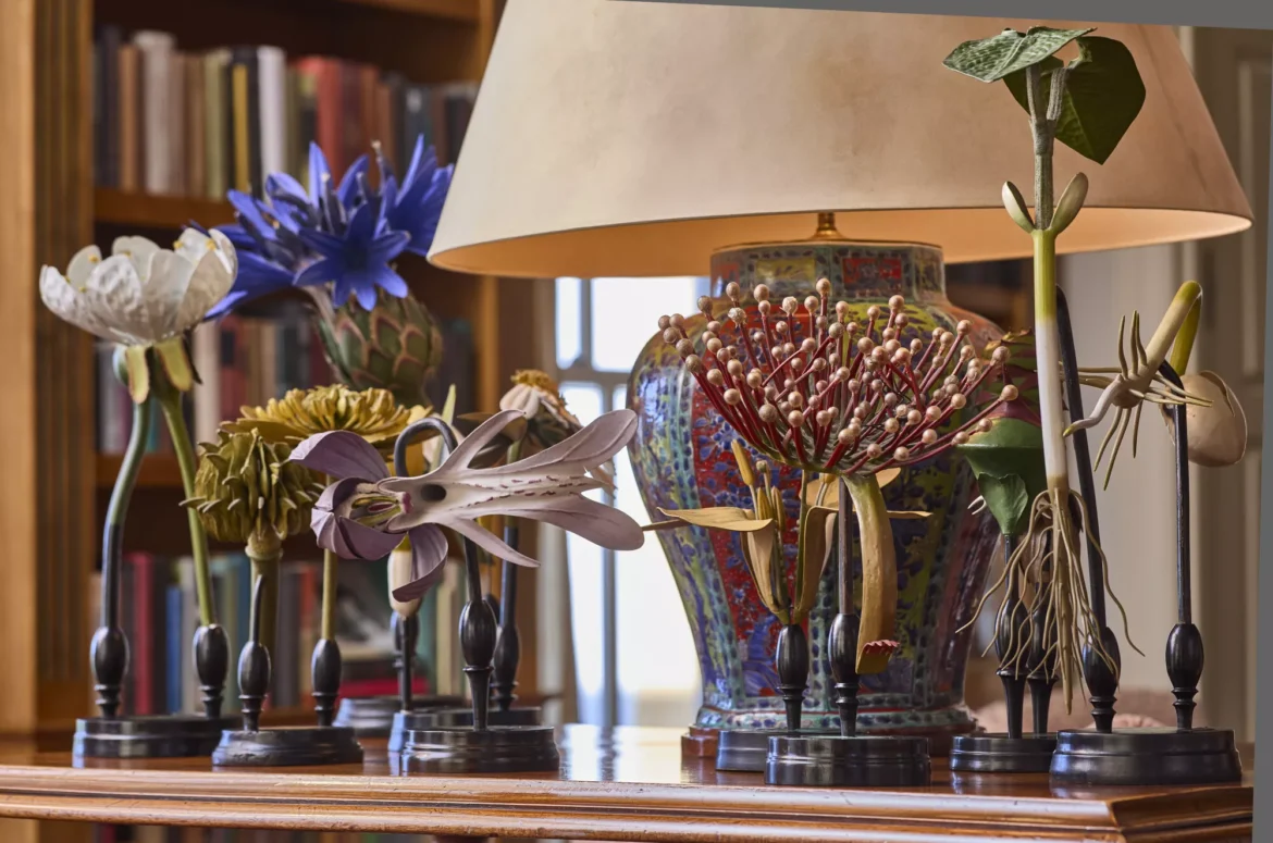 Intricate flower sculptures displayed on a wooden table, with a colorful vase and bookshelves in the background