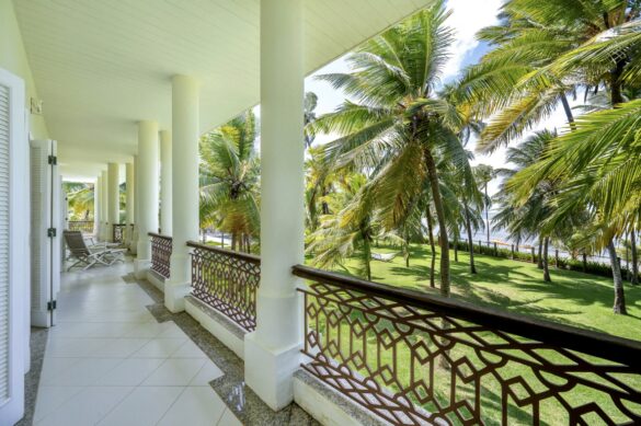 Bright bedroom suite opening to a columned veranda with ornate iron railings, featuring white linens and rattan furniture against a backdrop of swaying palms