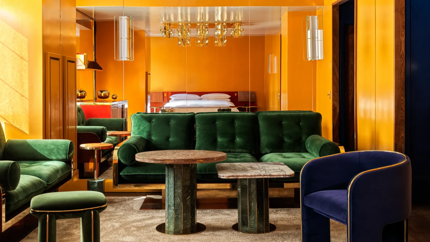 Luxury hotel guest room with orange walls, emerald green velvet sofa, marble side tables with green patinated bases, navy velvet chair with brass trim, and brass chandelier above bed