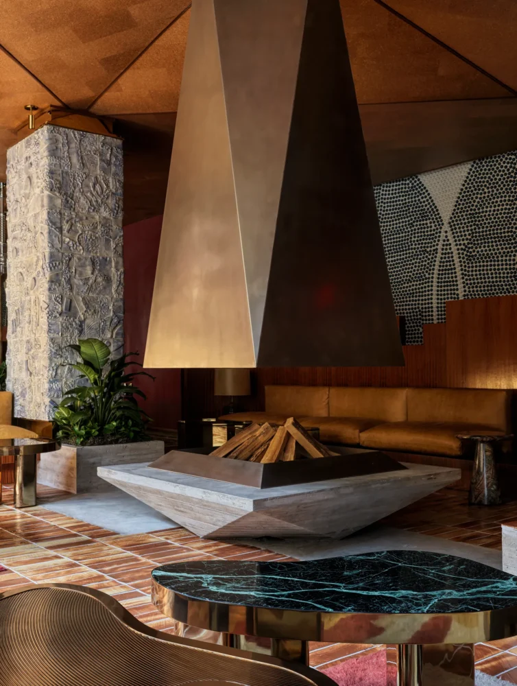 Modern hotel lounge with geometric metal hood fireplace, textured stone column, verde marble coffee table, and terracotta floor tiles